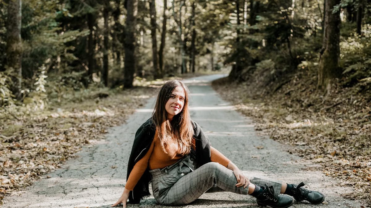 Cheerful young female tourist sitting on rough roadway between green trees in woods and looking at camera